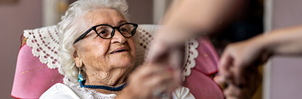 Female home carer supporting elder woman to stand up from an armchair