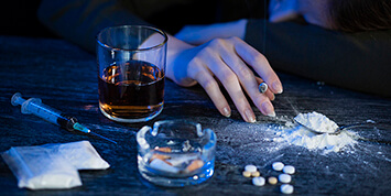 Woman's hand holding a lit cigarette resting on table next to glass of alcohol, ashtray, pills, and spilled baggies of powdered substance. 