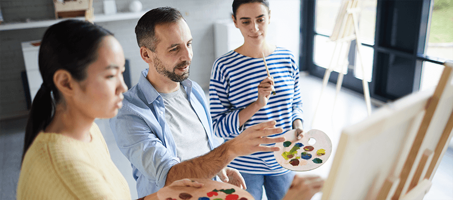 Group of people in art therapy holding paint palettes gathered around an easel.